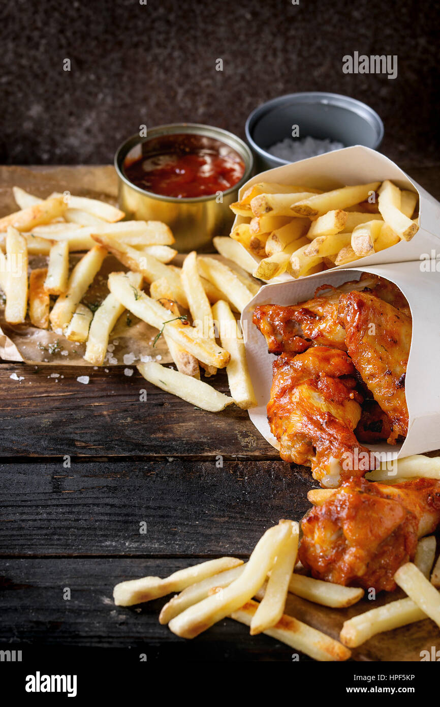 Fast food fried spicy chicken legs, wings and french fries potatoes in lunch boxes with salt and ketchup sauce served on baking paper over old dark wo Stock Photo