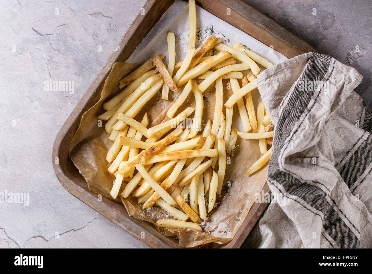 Fast food french fries potatoes with skin served with salt on baking paper in old rusty oven tray with kitchen towel over gray texture background. Top Stock Photo