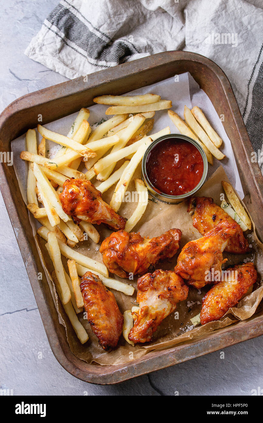Fast food fried spicy chicken legs, wings and french fries potatoes with salt and ketchup sauce served on baking paper in old oven tray with kitchen t Stock Photo