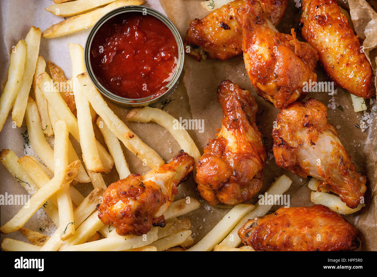 Fast food fried spicy chicken legs, wings and french fries potatoes with salt and ketchup sauce served on baking paper. Top view, close up Stock Photo