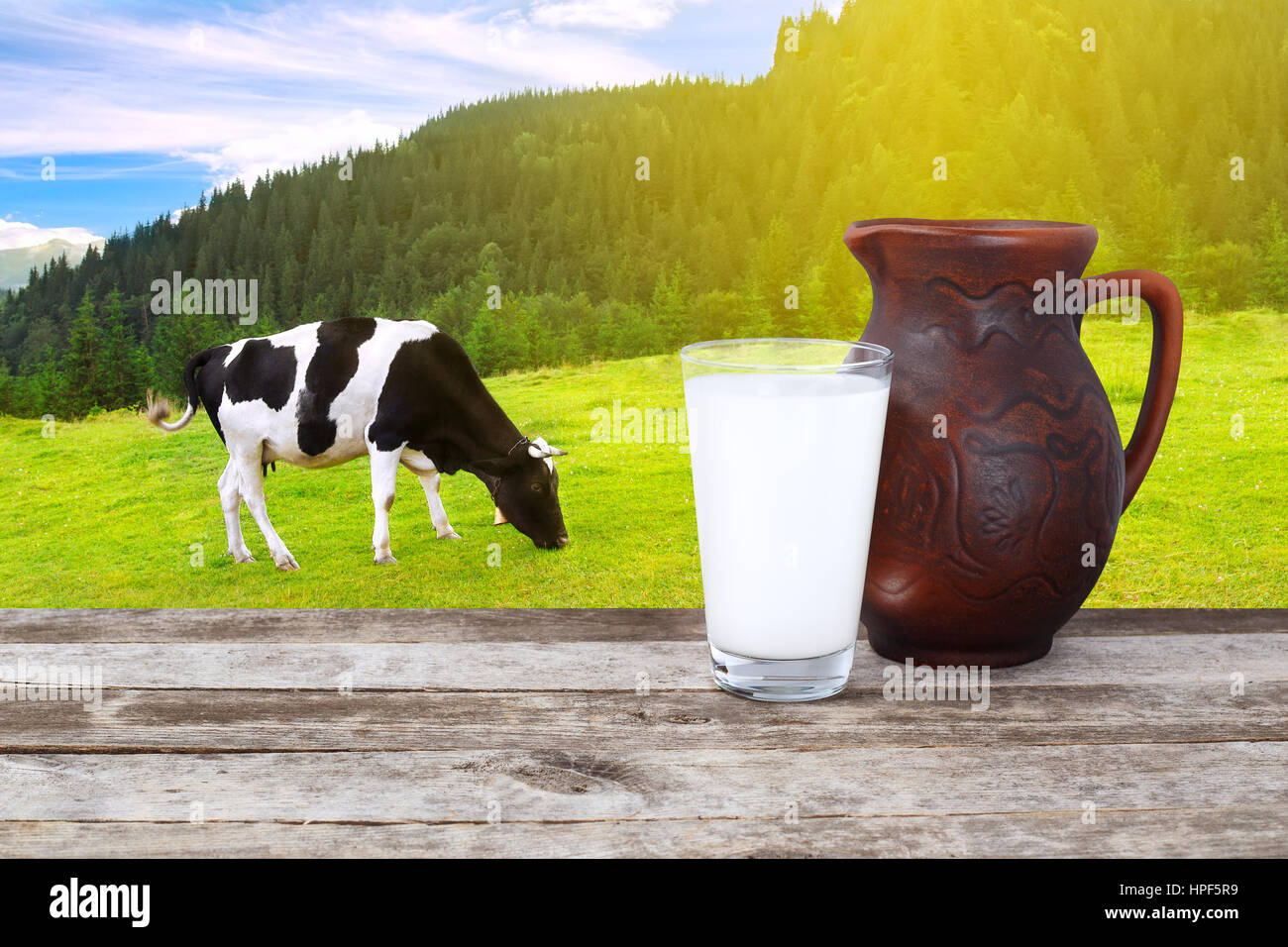 Milk in glass and clay jug on old wooden table with grazing cow on the ...
