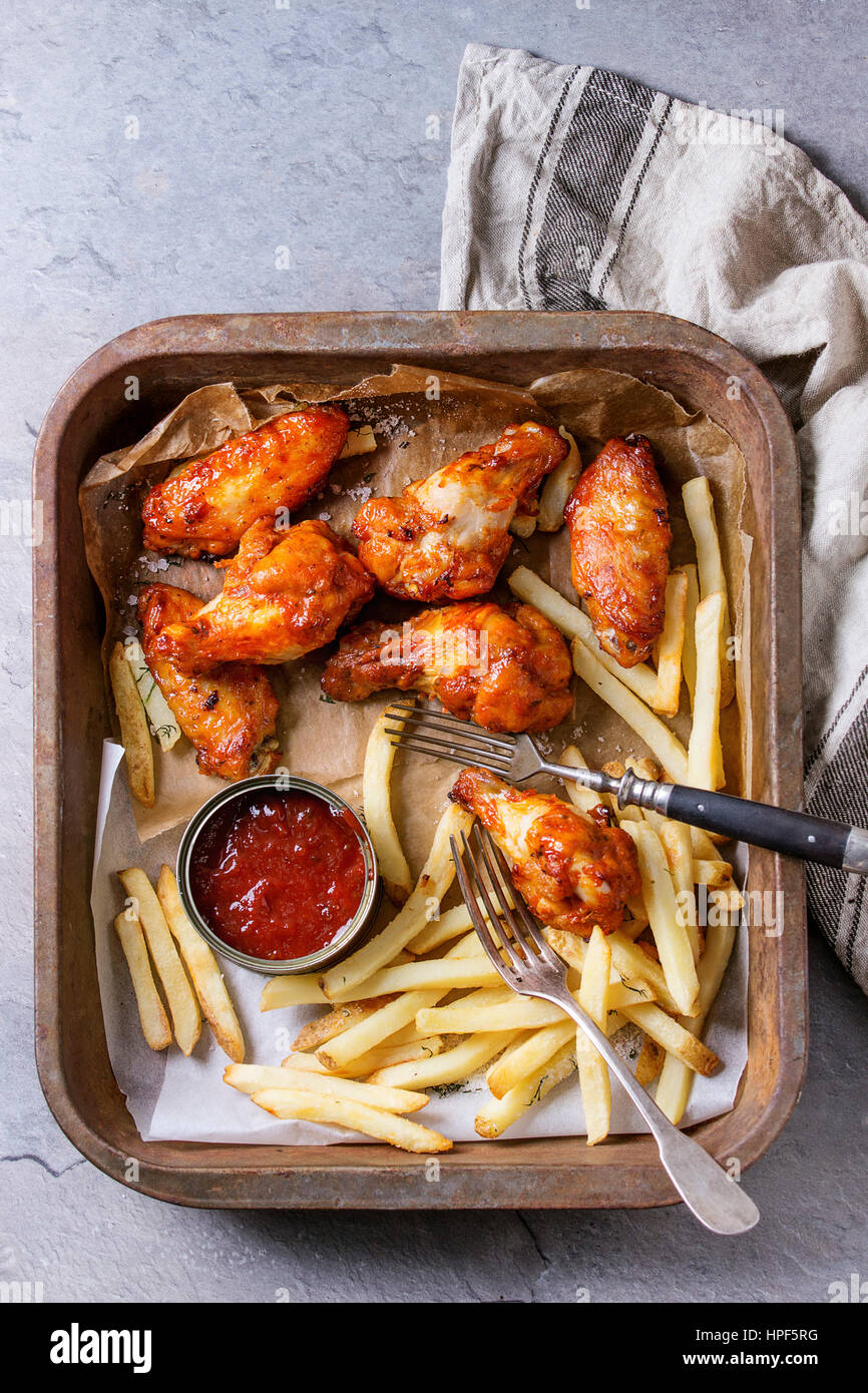 Fast food fried spicy chicken legs, wings and french fries potatoes with salt and ketchup served on baking paper in old oven tray with kitchen towel o Stock Photo