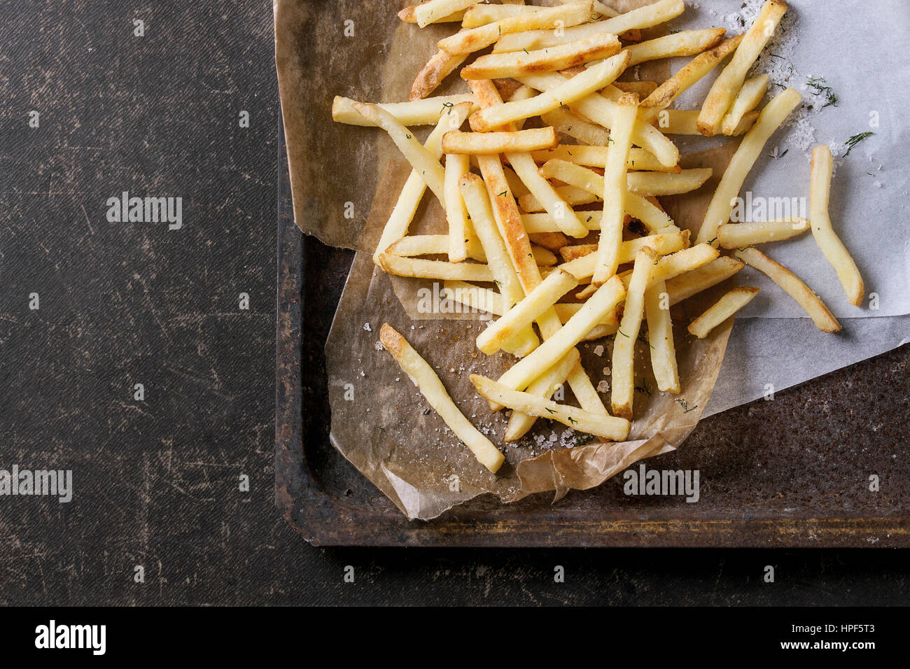 Fast food french fries potatoes with skin served with salt and herbs on baking paper on old rusty oven tray over dark texture background. Top view, sp Stock Photo