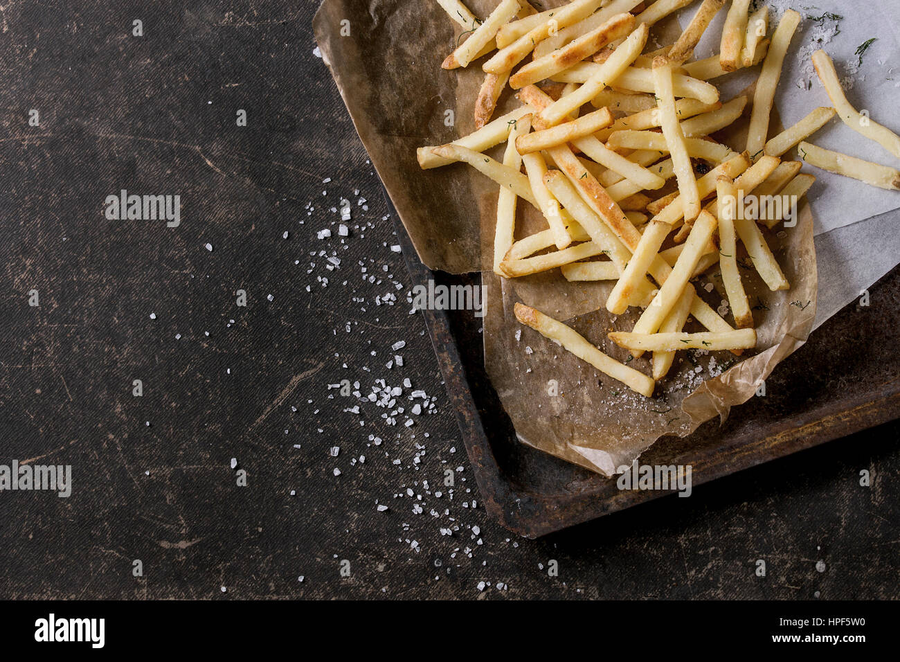 Fast food french fries potatoes with skin served with salt and herbs on baking paper on old rusty oven tray over dark texture background. Top view, sp Stock Photo