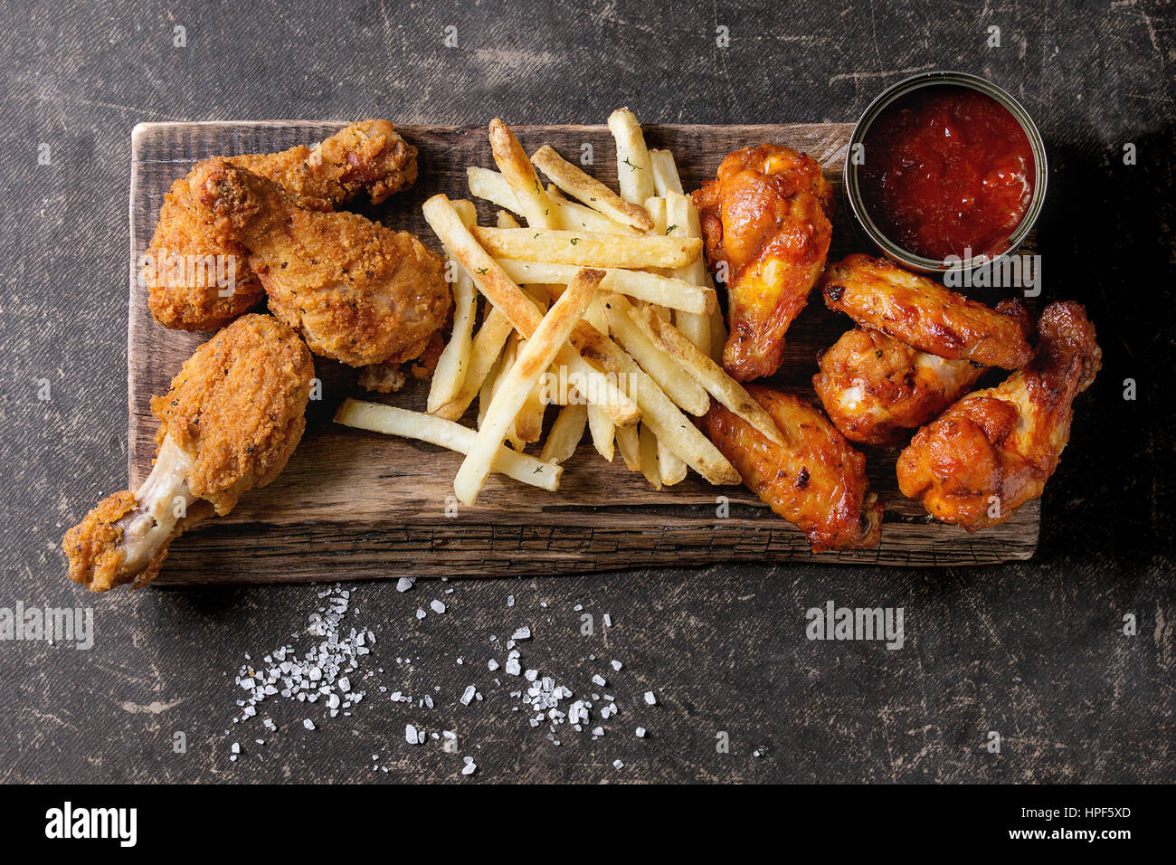 Fast food fried crispy and spicy chicken legs, wings and french fries potatoes with salt and ketchup sauce served on wooden serving board over dark te Stock Photo