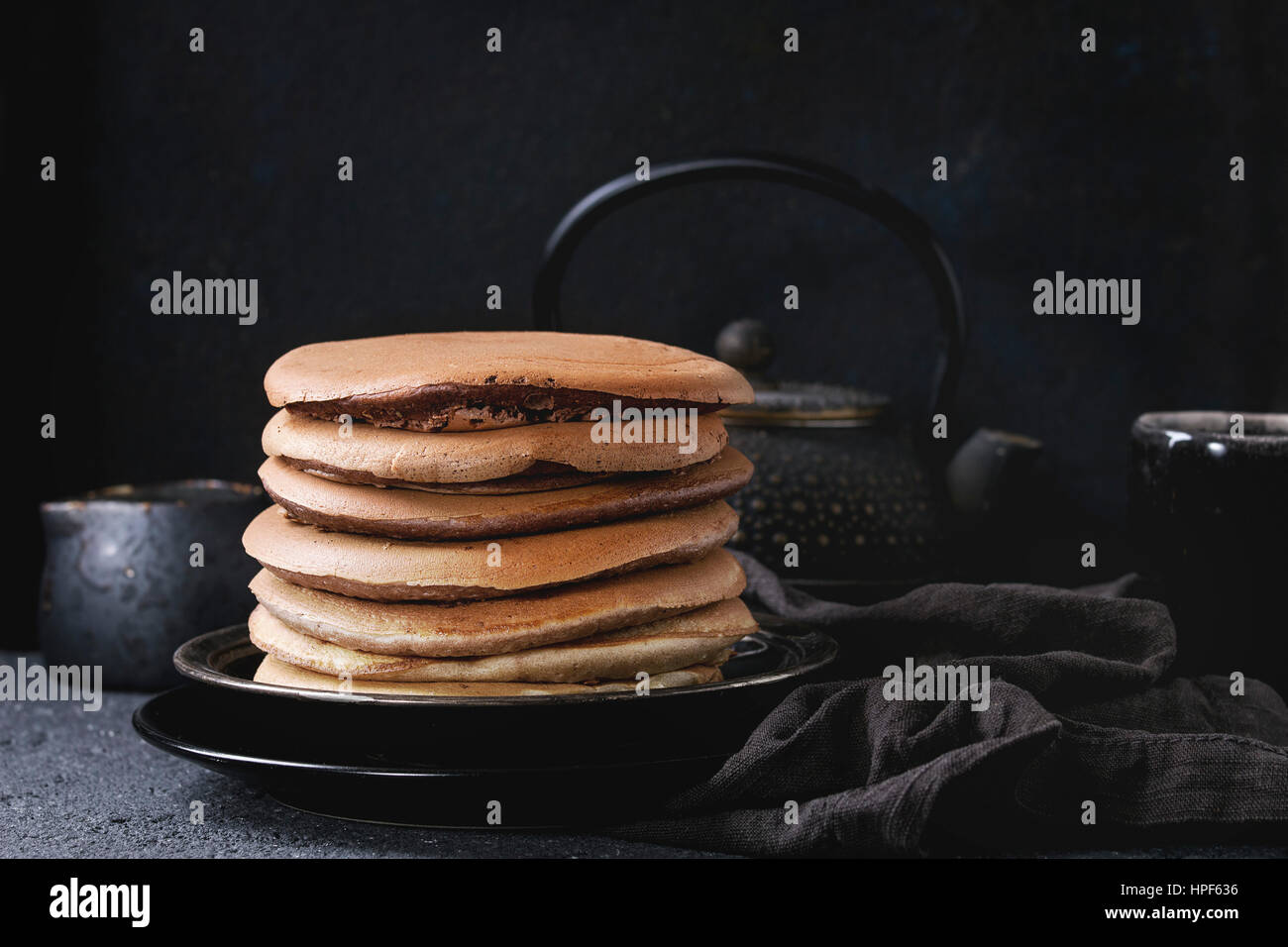 Stack of homemade american ombre chocolate pancakes served on black plate with jug of cream and teapot over black stone texture background. Stock Photo