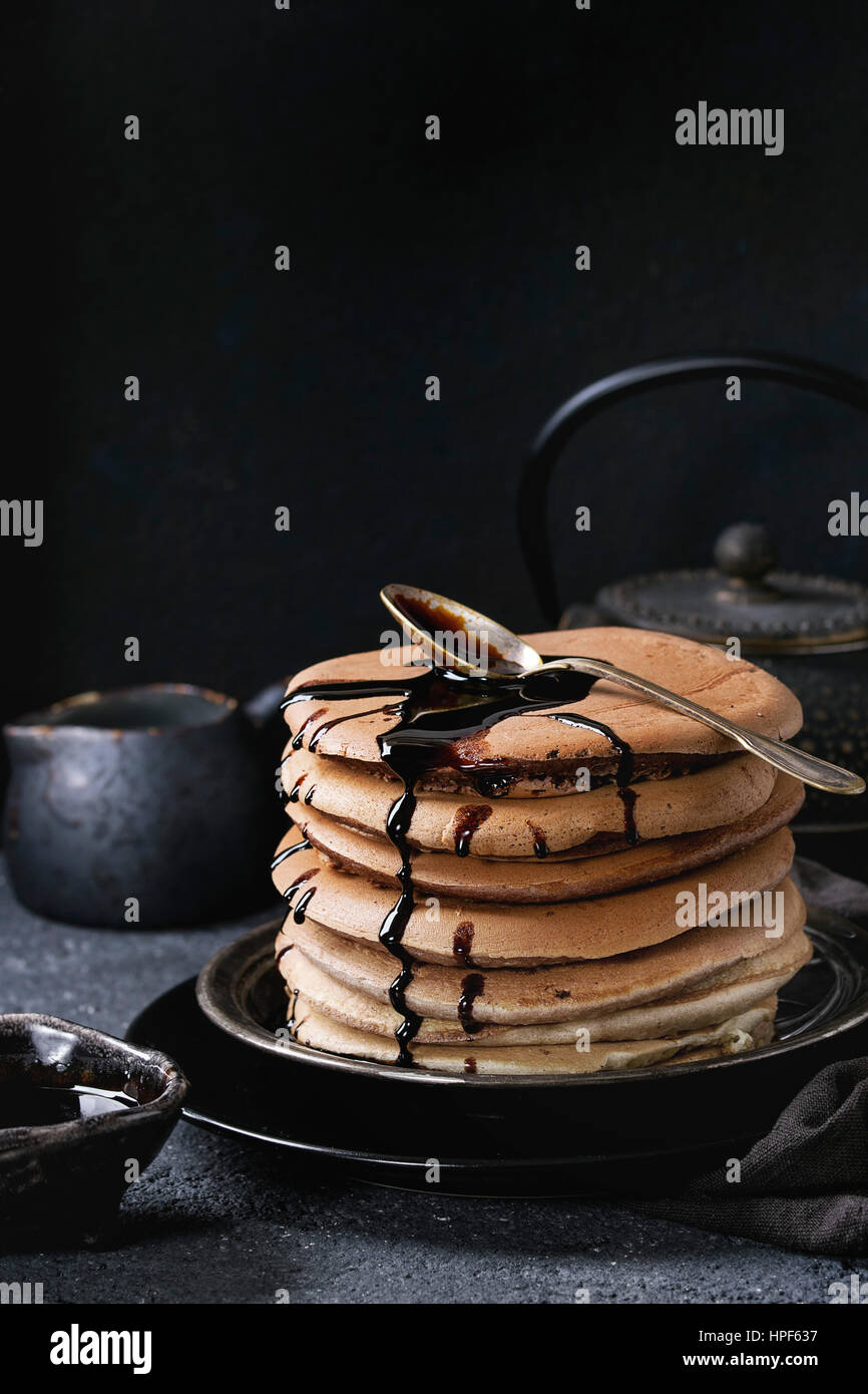 Stack of homemade american ombre chocolate pancakes with carob honey sauce served on black plate with jug of cream and teapot over black stone texture Stock Photo