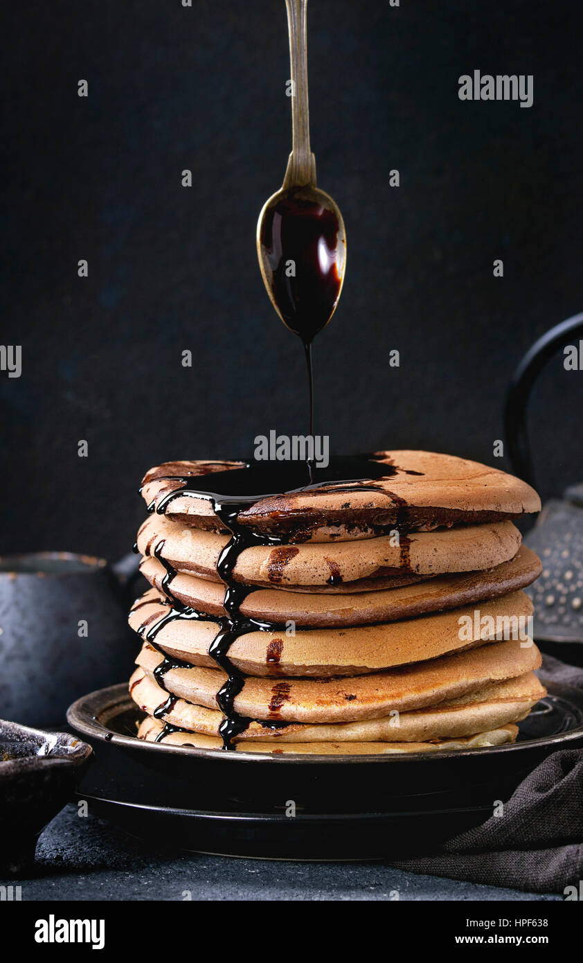 Stack of homemade american ombre chocolate pancakes with carob honey sauce, flowing from spoon, served on black plate with jug of cream and teapot ove Stock Photo