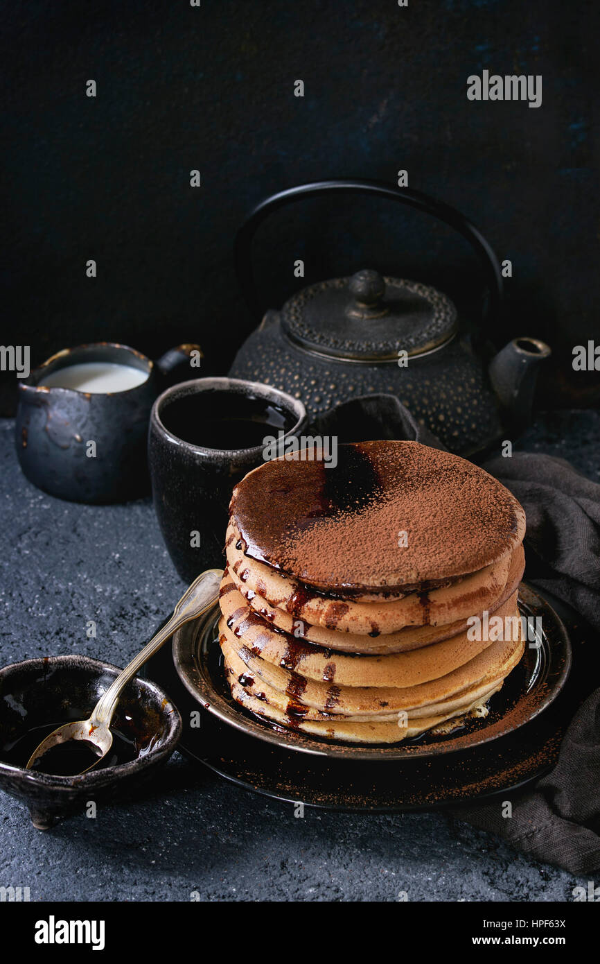 Stack of homemade american ombre chocolate pancakes with carob honey sauce and cocoa powder served on black plate with jug of cream and teapot over bl Stock Photo