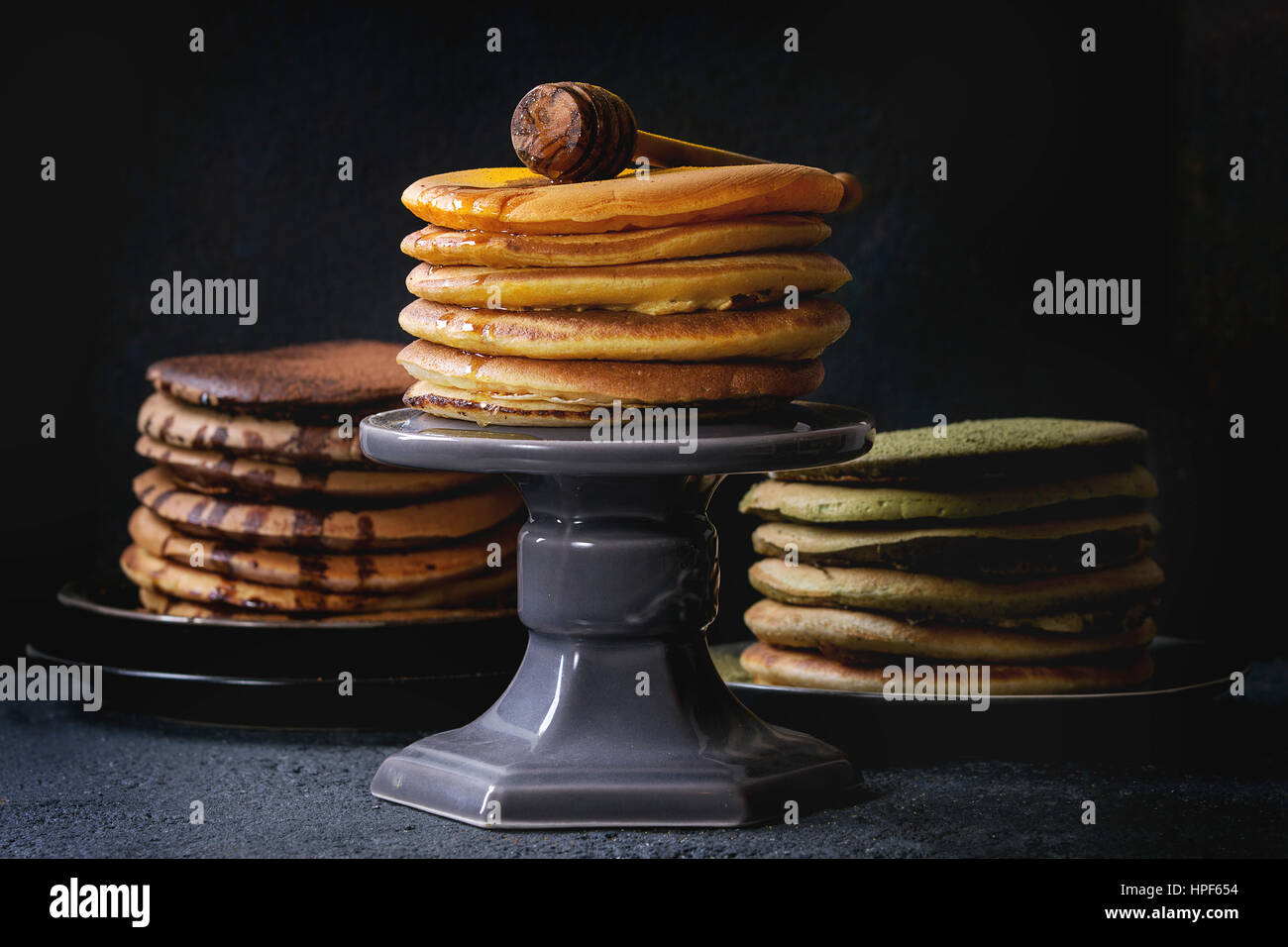 variety of homemade american ombre chocolate, green tea matcha and turmeric pancakes with honey and sauce served on plates and cake stand over black s Stock Photo