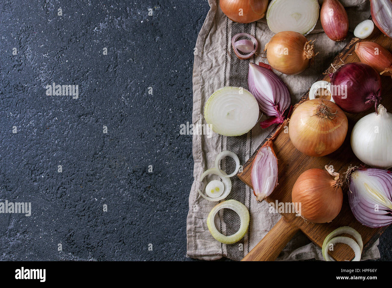 Variety of whole and sliced red, white, yellow and shallot onions on wooden chopping cutting board on textile napkin over dark stone texture backgroun Stock Photo