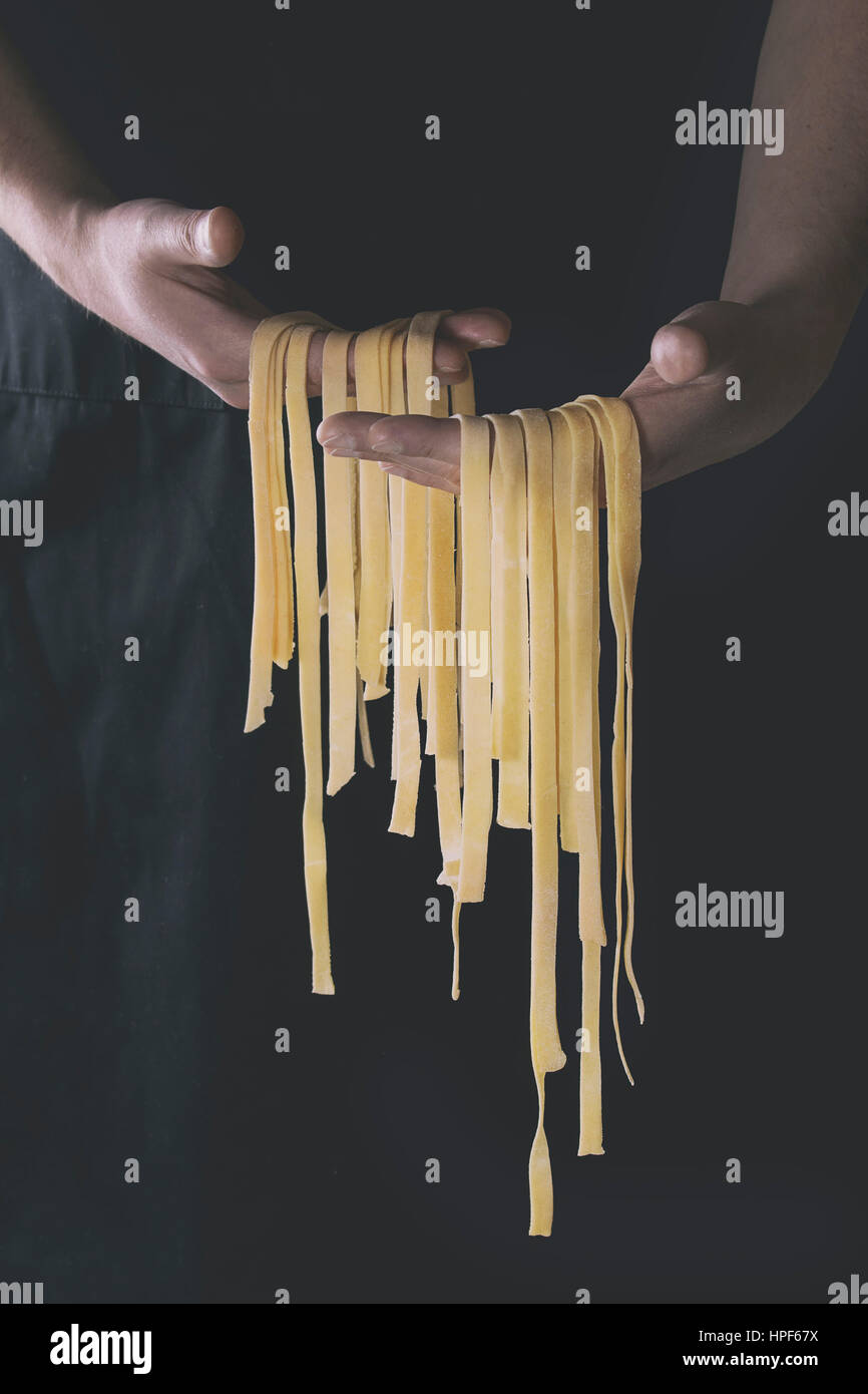 Fresh raw uncooked homemade pasta tagliatelle in man's hands over black apron as background. Retro filter toned Stock Photo