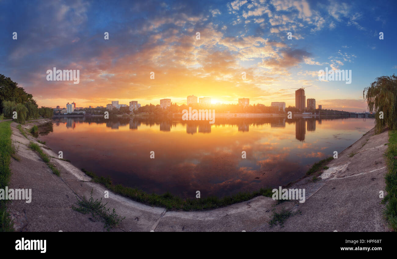 Beautiful cityscape at sunset. Amazing panoramic view of the city from the river bank. Colorful blue sky with clouds and sun reflected in water Stock Photo