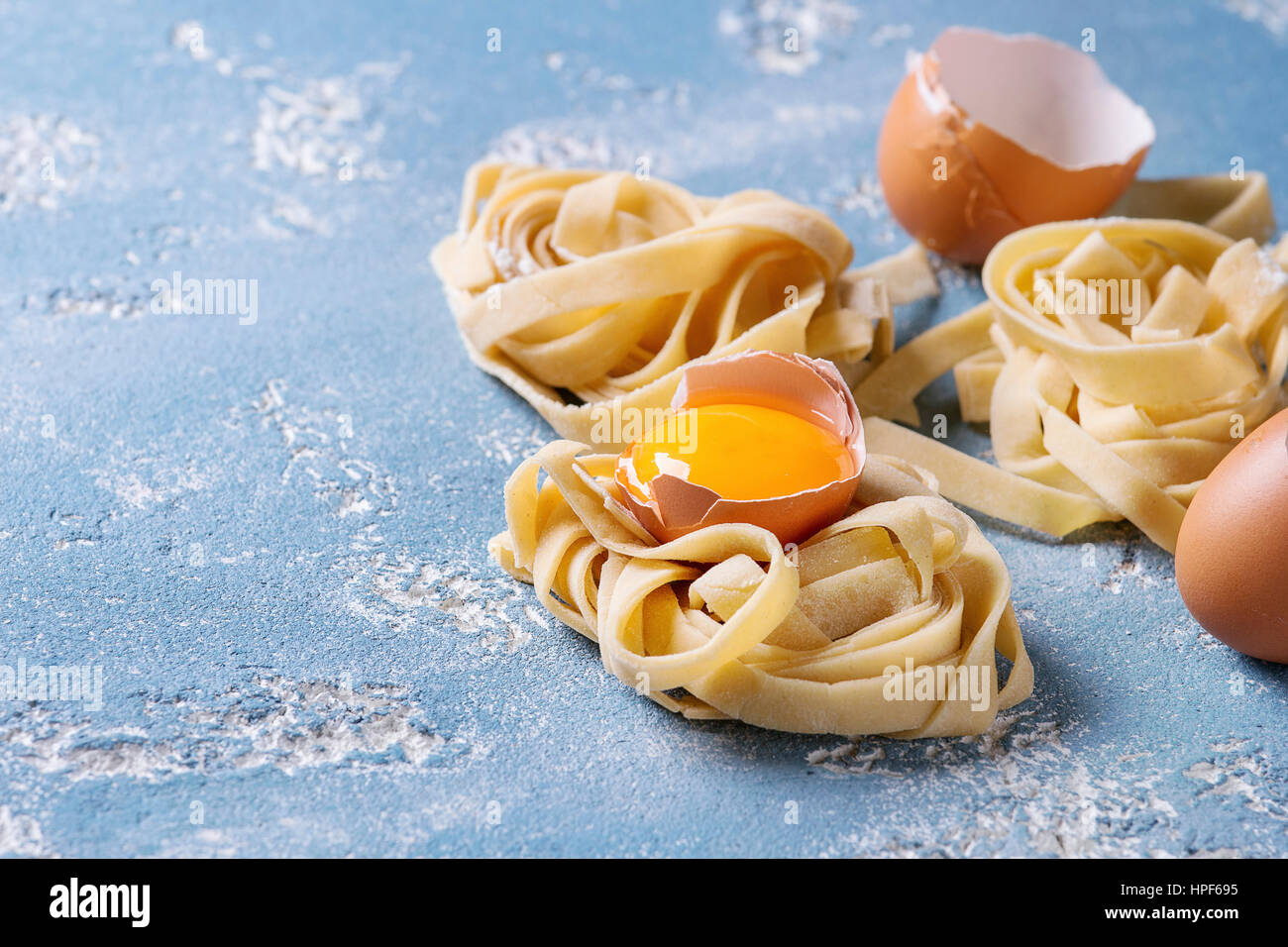 Fresh raw uncooked homemade twisted pasta tagliatelle with egg yolk, shell and pasta cutter over light blue concrete background. Stock Photo