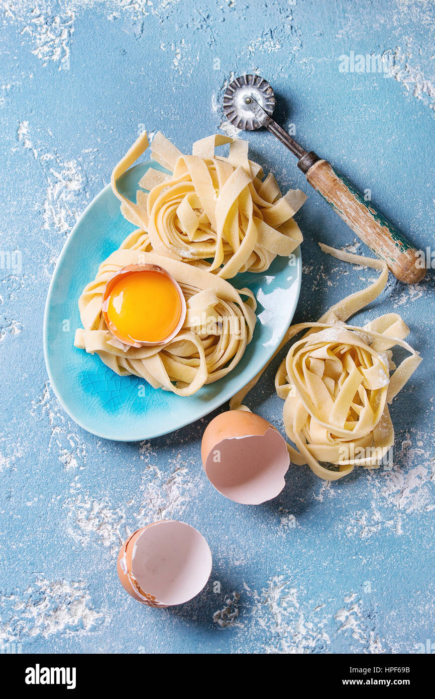Fresh raw uncooked homemade twisted pasta tagliatelle with egg yolk, shell and pasta cutter in wooden tray with kitchen towel over light blue concrete Stock Photo