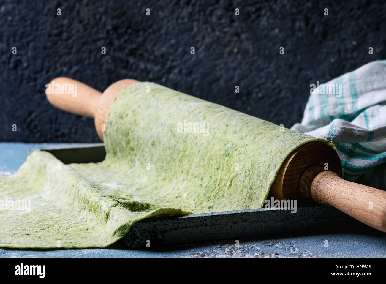 Fresh homemade green spinach rolled dough for pasta tagliatelle on wood rolling pin with egg yolk and kitchen towel over dark gray background. Top vie Stock Photo