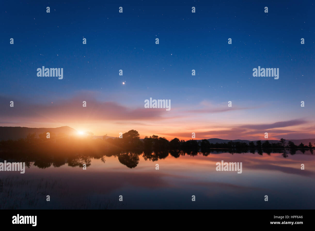 Mountain lake with moonrise at night. Night landscape with river, trees, hills, moon, stars and colorful blue sky with clouds reflected in water Stock Photo