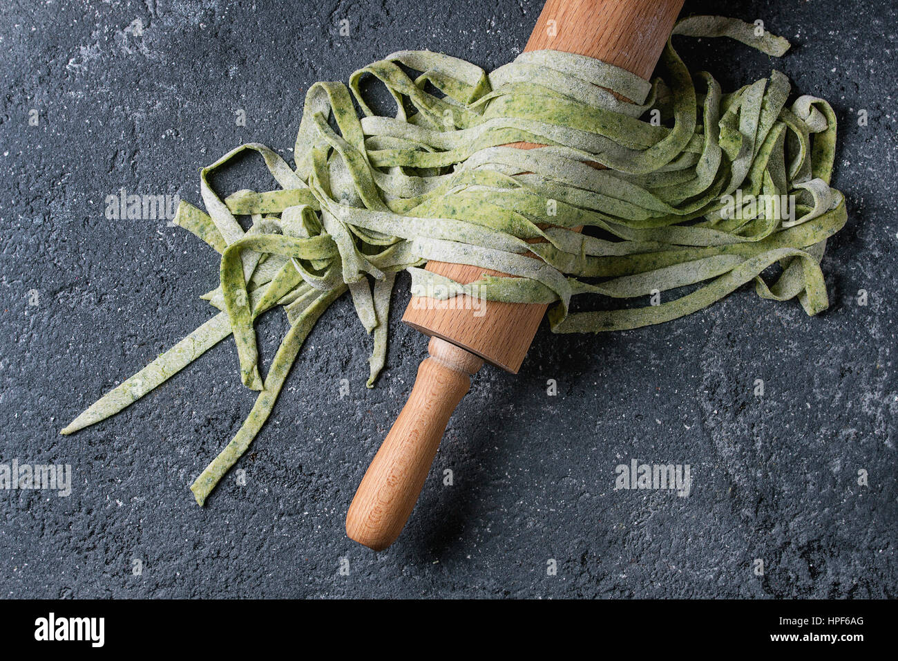 Fresh raw uncooked homemade green spinach pasta tagliatelle on wooden rolling pin over dark texture concrete background. Top view with space. Stock Photo