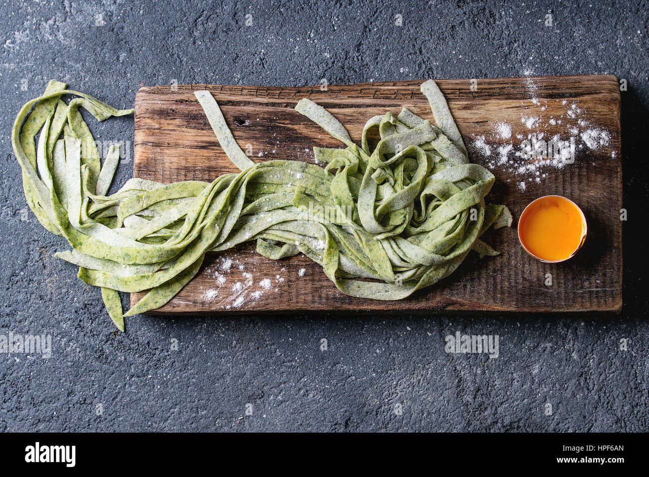 Fresh raw uncooked homemade green spinach pasta tagliatelle with egg yolk and flour on wooden chopping cutting board over dark texture concrete backgr Stock Photo