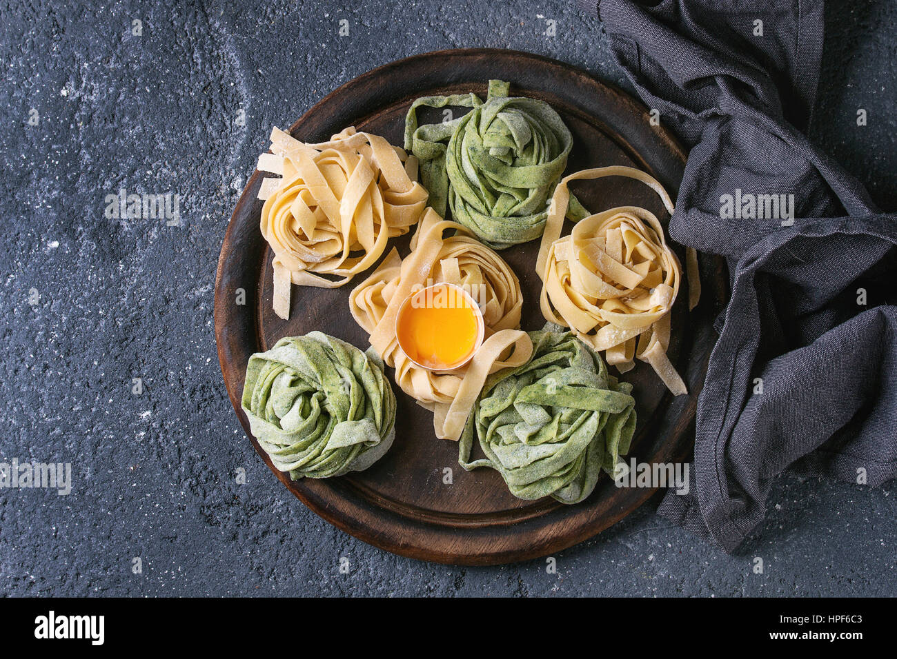 Variety of colored fresh raw uncooked homemade pasta tagliatelle green spinach and traditional yellow with egg yolk on wooden cutting board over dark Stock Photo