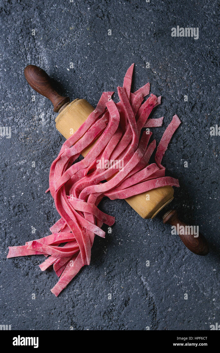 Fresh raw uncooked homemade pink beetroot pasta tagliatelle on wooden rolling pin over dark texture concrete background. Top view with space. Stock Photo