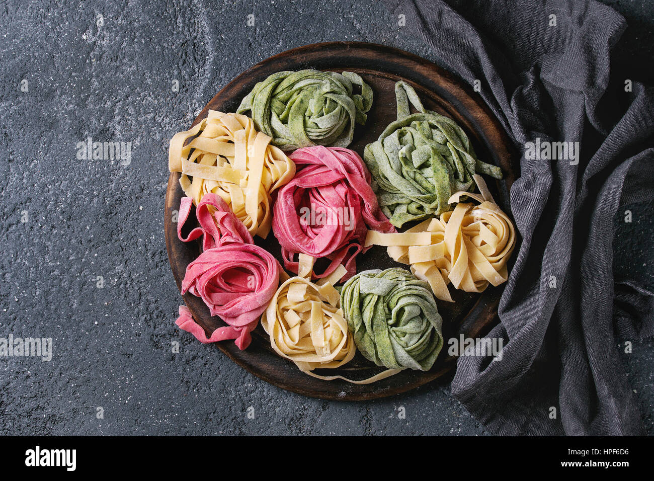 Variety of colored fresh raw uncooked homemade pasta tagliatelle green spinach, pink beetroot and yellow on wooden cutting board with textile over dar Stock Photo