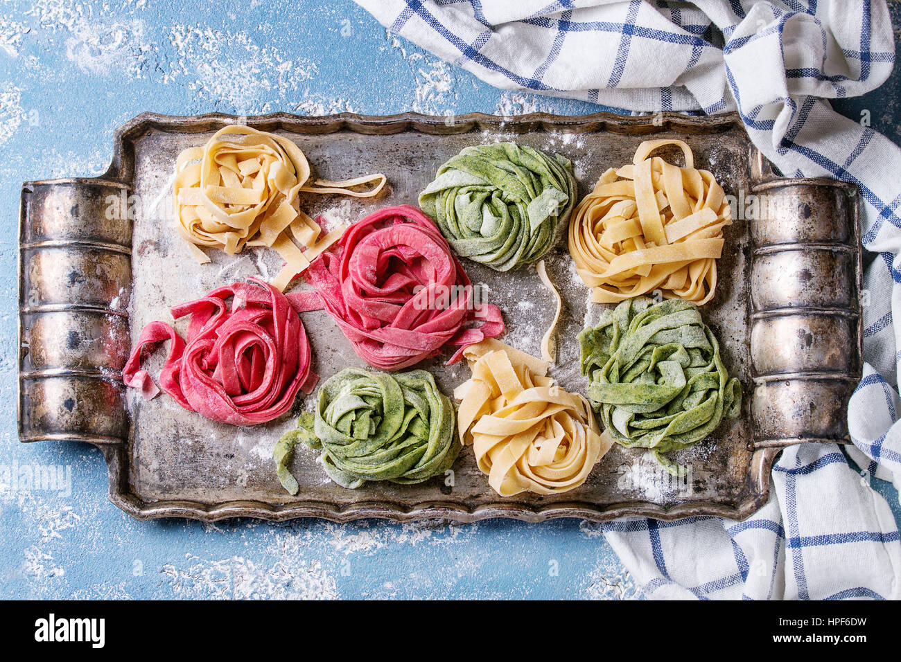 Variety of colored fresh raw uncooked homemade pasta tagliatelle green spinach, pink beetroot and yellow with flour on old metal tray over blue concre Stock Photo