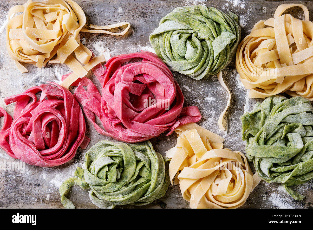 Variety of colored fresh raw uncooked homemade pasta tagliatelle green spinach, pink beetroot and yellow with flour over old metal texture background. Stock Photo