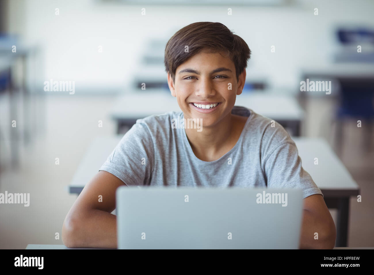 Portrait of happy schoolboy sitting with laptop in classroom at school ...