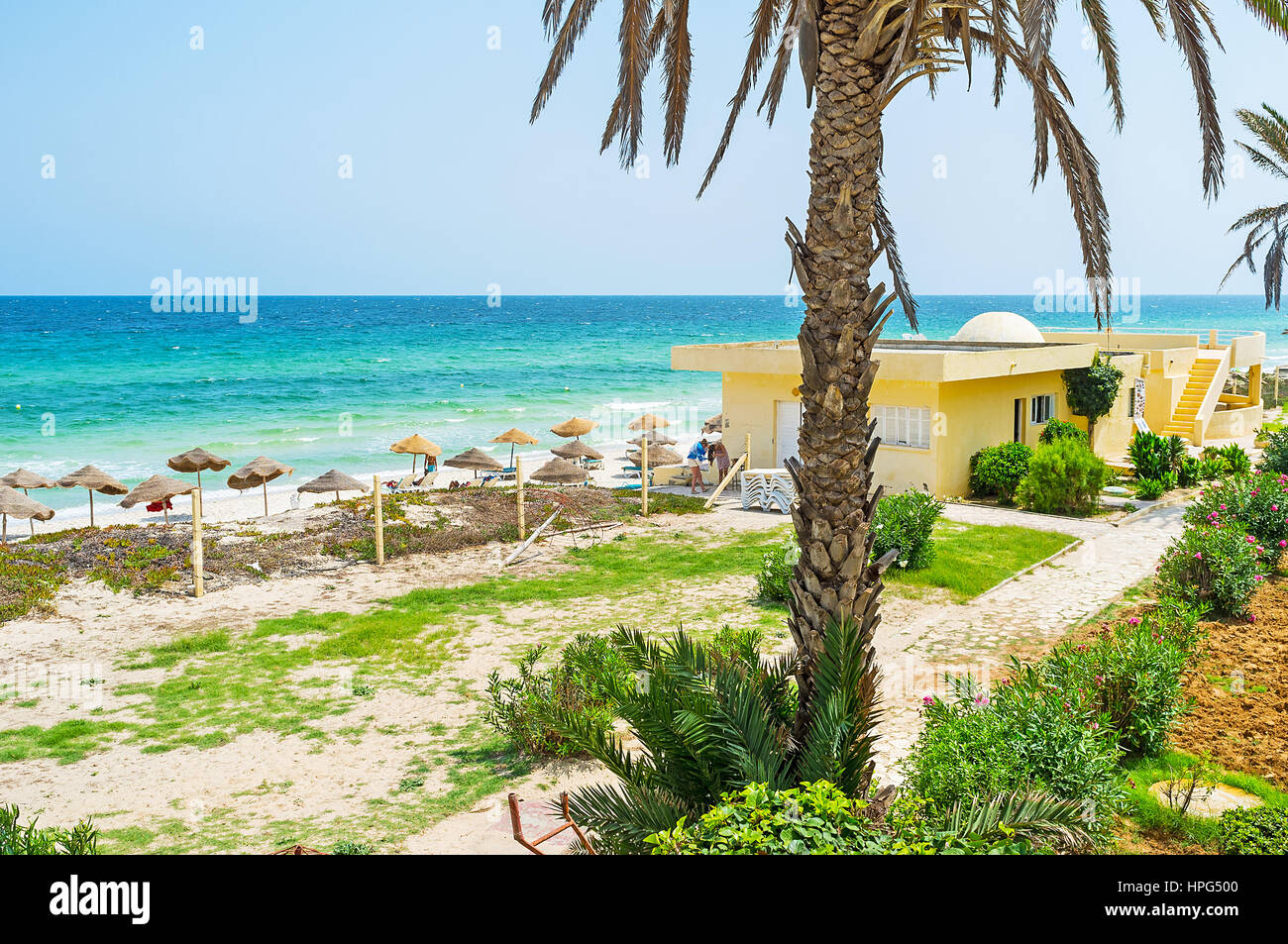 The cozy beach with straw sunshades neighbors with the scenic green garden with palms and blooming bushes, El Kantaoui resort, Tunisia. Stock Photo