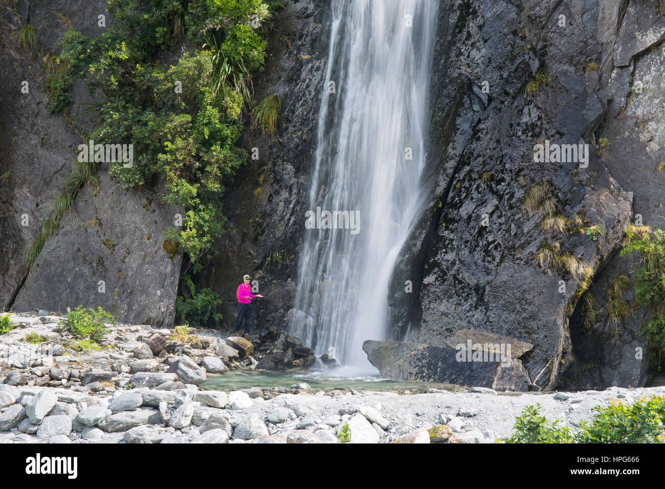 Franz Josef, Westland Tai Poutini National Park, West Coast, New Zealand. Visitor posing beneath waterfall on the Franz Josef Glacier Valley Track. Stock Photo