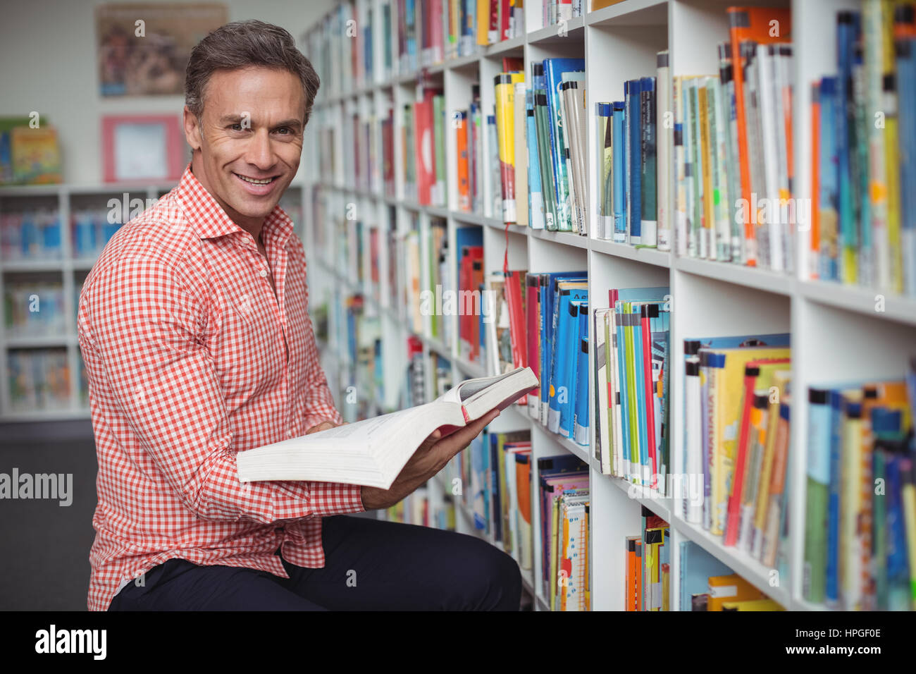 Portrait of smiling school teacher holding book in library at school ...
