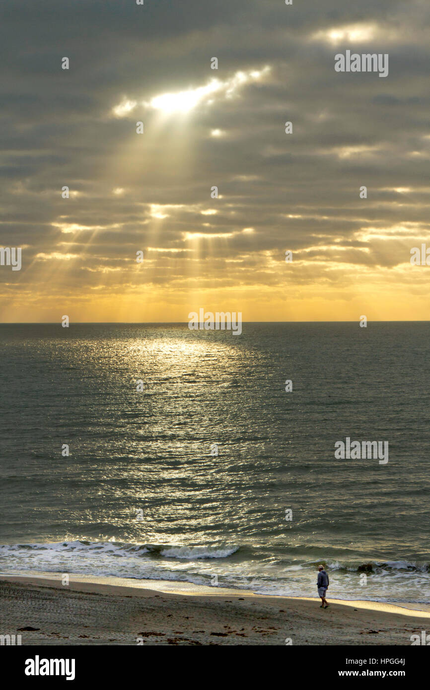 Light rays pour down breaking through dark clouds onto a calm sea iluminating a luminous circle of light as a lone man walks the beach Stock Photo