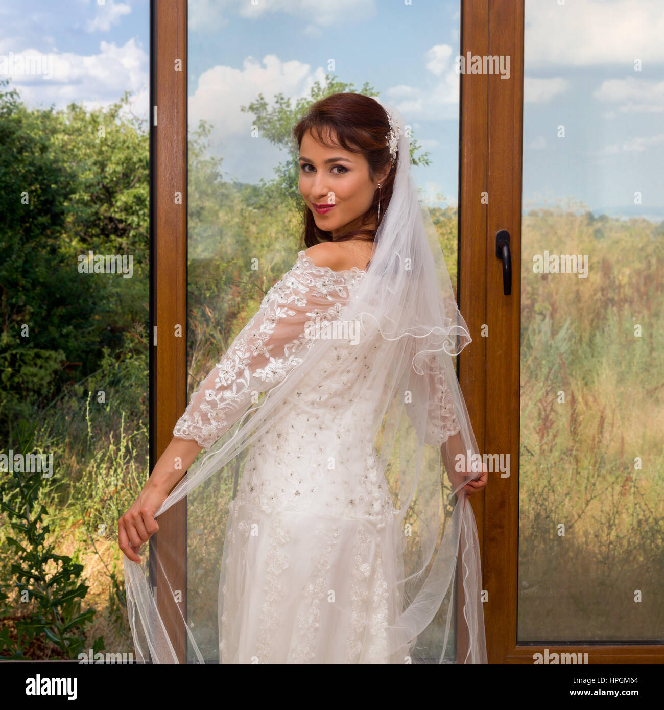 Young bride in traditional wedding dress standing in front of sliding doors or window Stock Photo