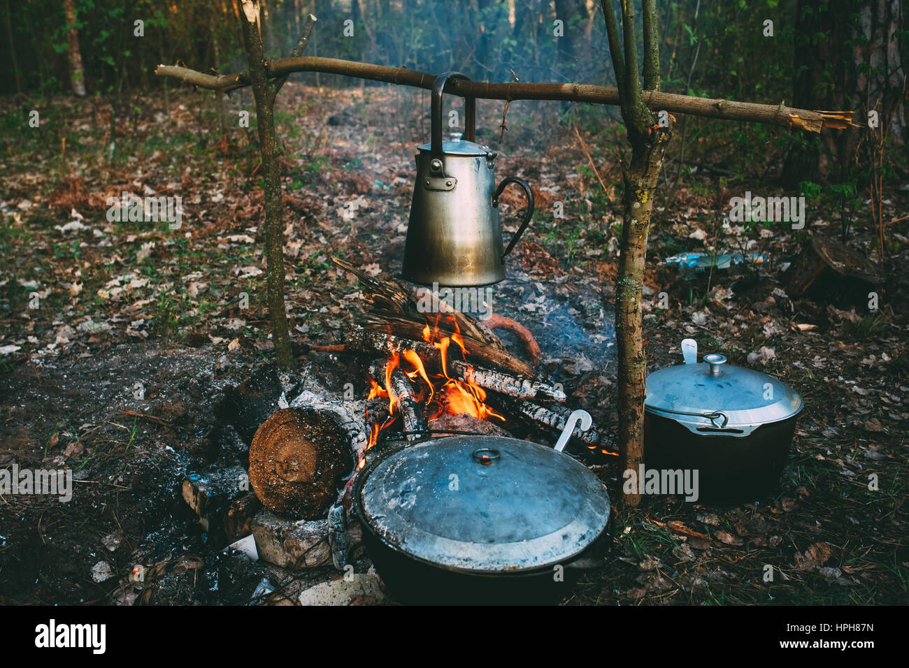 Old Retro Iron Camp Kettle And Pans Boils Water On A Fire In Forest. Bright Flame Fire Bonfire At Dusk Night. Stock Photo
