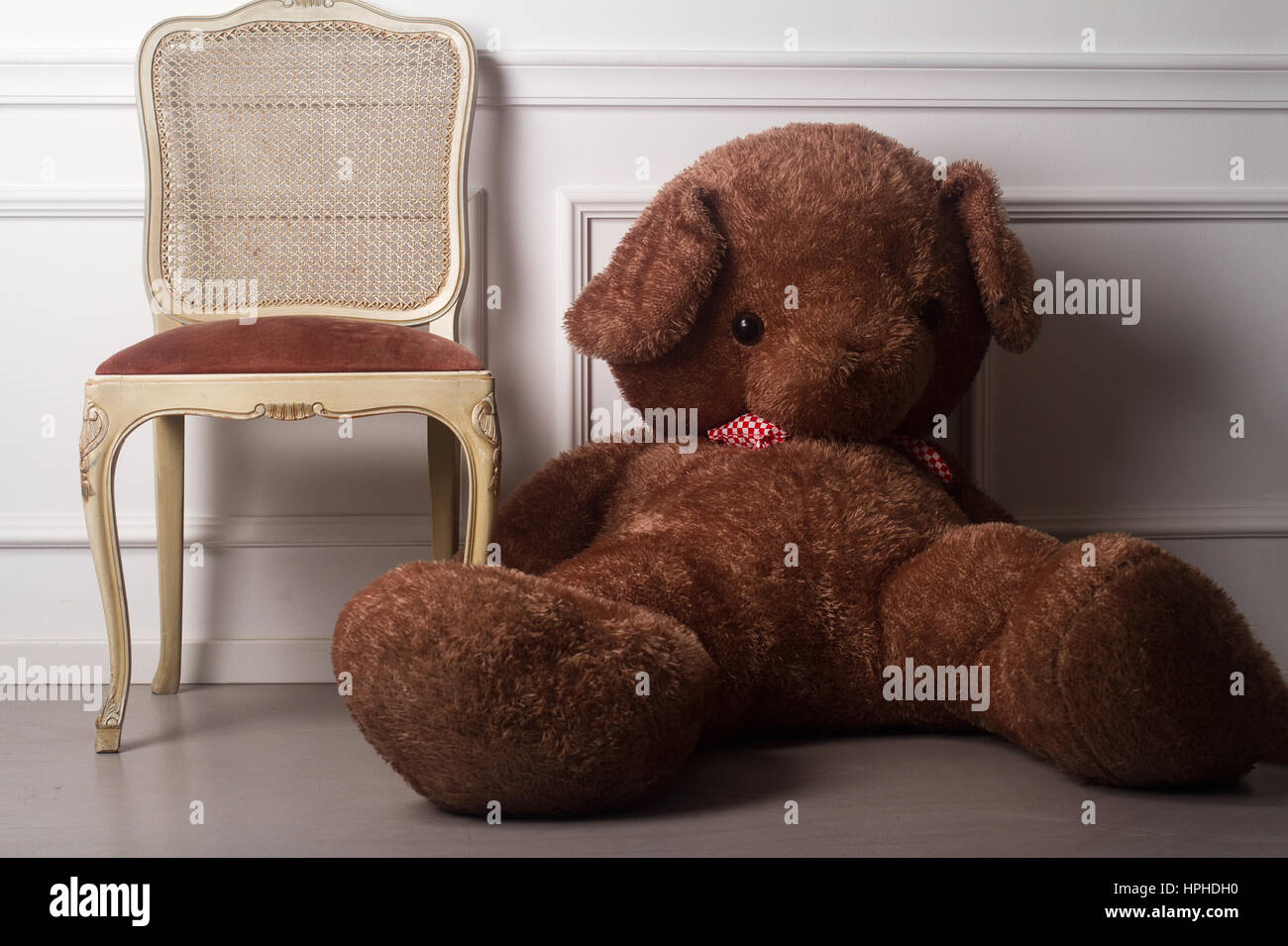 horizontal front view of a large brown teddy bear sitting on the floor next to a luxury chair against a white wall Stock Photo