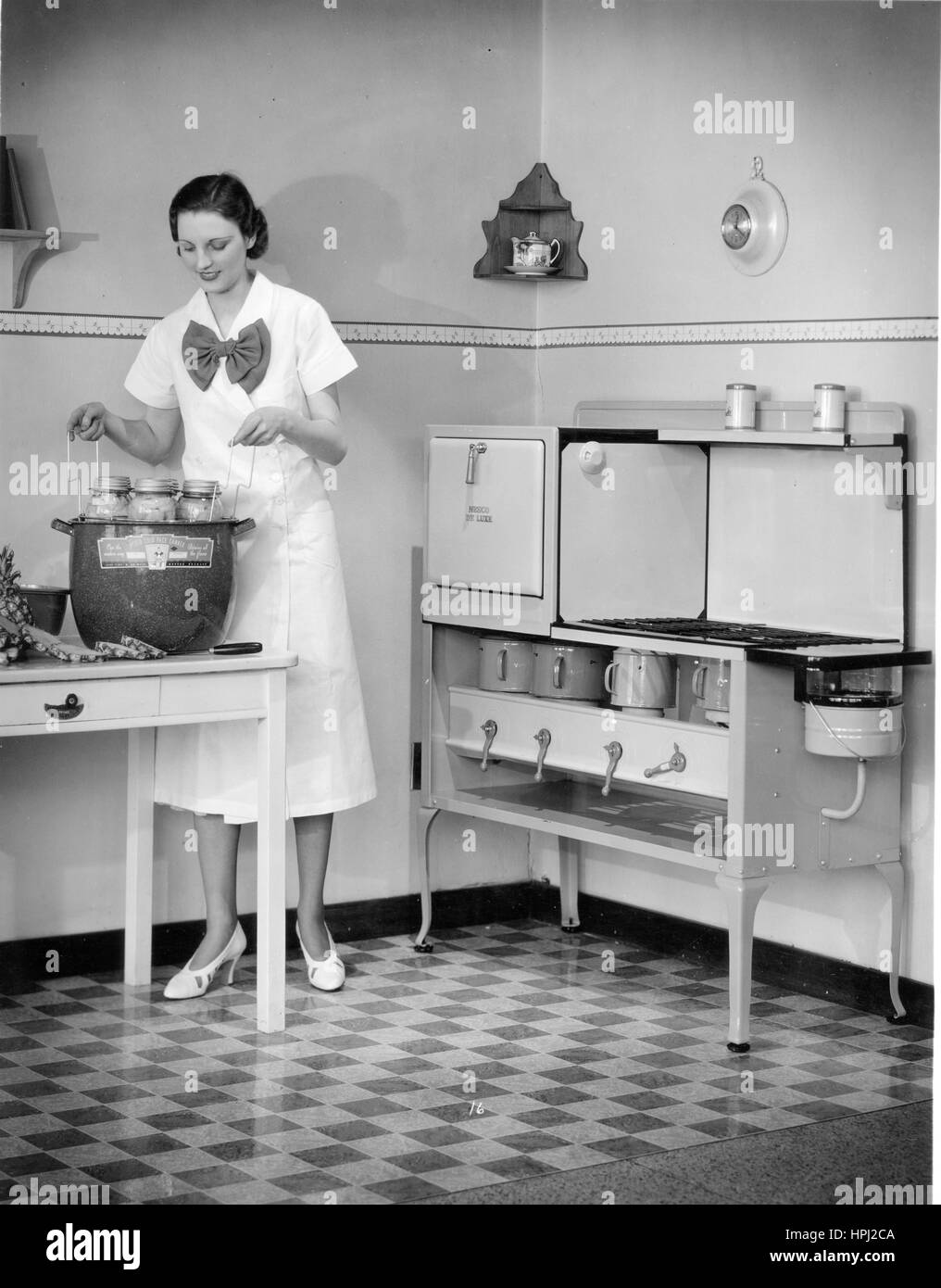 A prim homemaker in front of a 'NESCO De Luxe' oil cook stove in a 1920s kitchen.  She is canning fruits (in glass jars), using her 'NESCO Cold Pack Canner,' that holds 6 jars at once.  Sliced Pineapples lay on the table.   To see my related vintage images, Search:  Prestor  vintage  woman Stock Photo