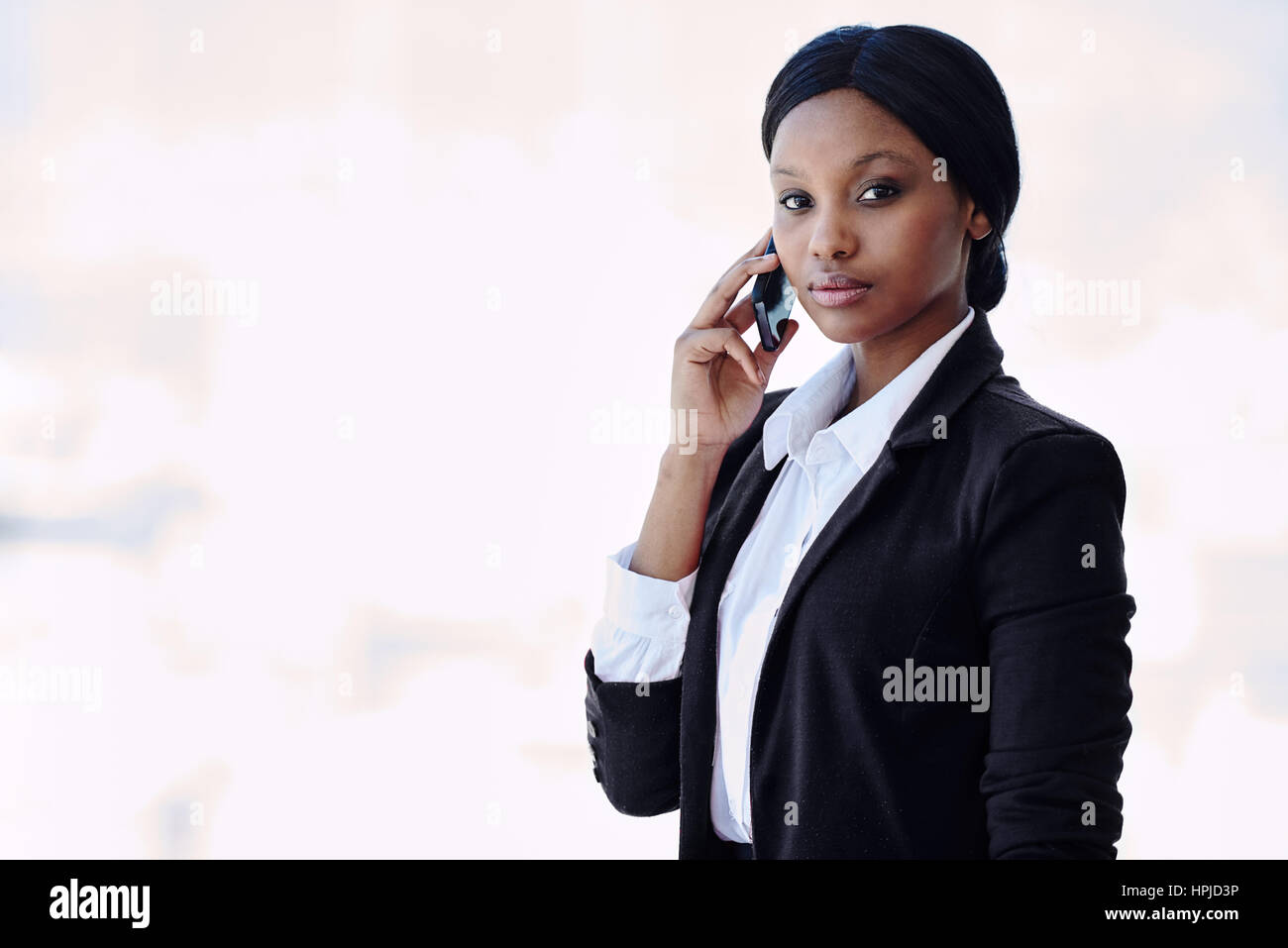 Young attractive, well dressed black businesswoman looking at camera with a serious facial expression while holding her phone against her ear in her r Stock Photo