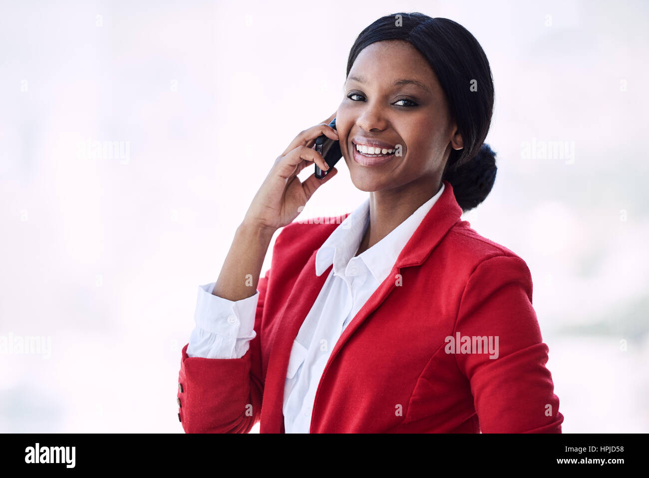 Attractive young black businesswoman smiling at the camera while talking on her mobile phone that she is holding in her right hand, all while looking  Stock Photo