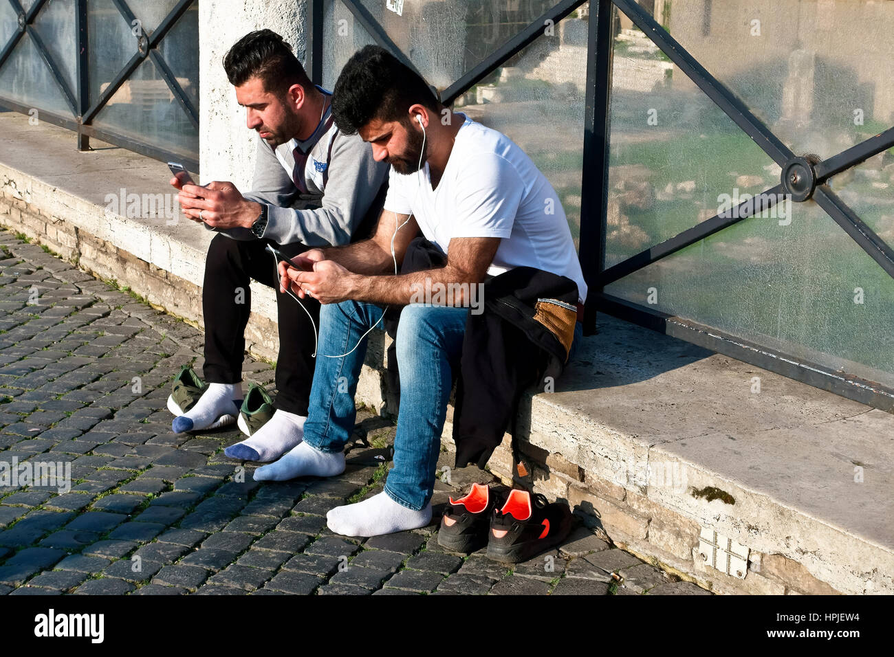 Two tourists friends using their smartphones, relaxing with their shoes off while visiting the Trajan’s roman forum. Rome, Latium, Italy, Europe, EU. Stock Photo
