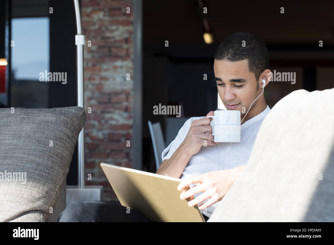 man using a digital tablet at home on his sofa in his living room, while listening to music and drinking a hot coffee or tea Stock Photo