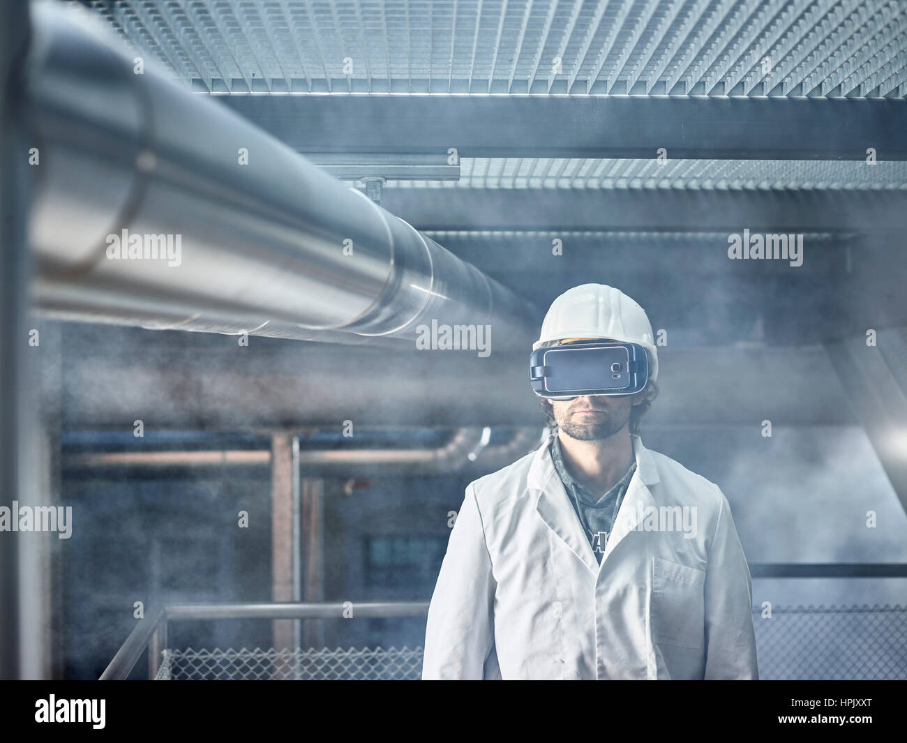 Man with VR goggles, white helmet and lab coat standing in front of industrial plant, Austria Stock Photo