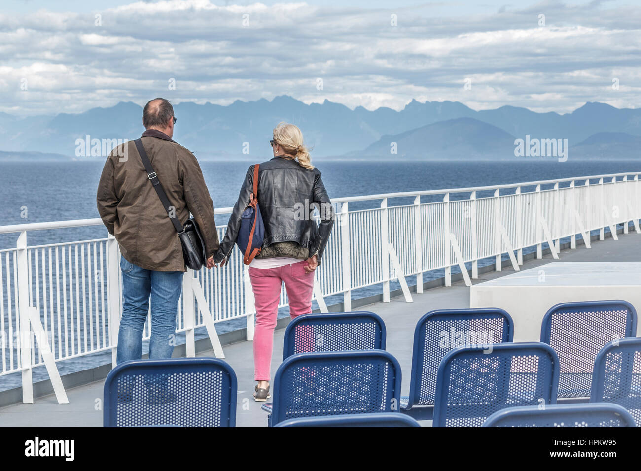 A couple, alone on the deck of a ferry, hold hands and look across the Strait of Georgia to Bowen Island, Howe Sound and BC's Coast Mountain Range. Stock Photo