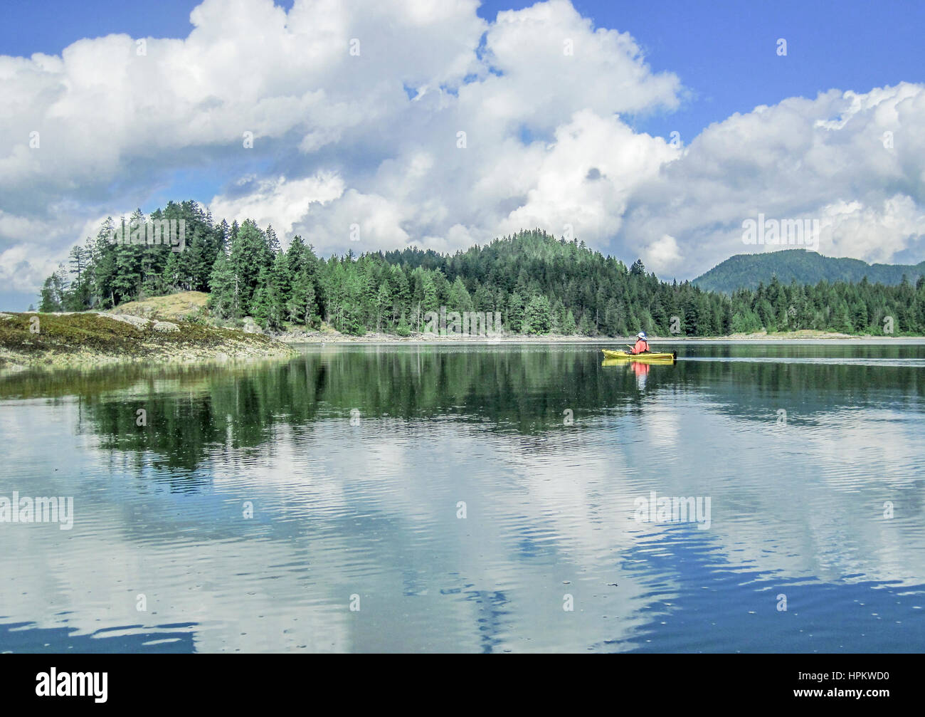 A 59 year old man kayaks beside a forested island in British Columbia, with towering clouds reflected in the water and a light rain falling. Stock Photo
