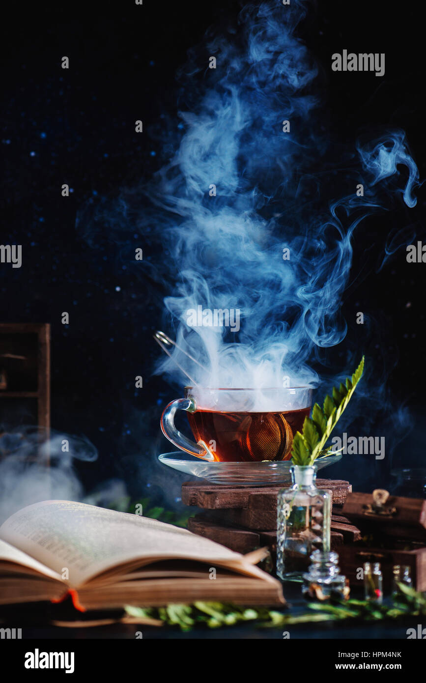 A cup of tea with a dense steam, an open book, glass bottles and herbs