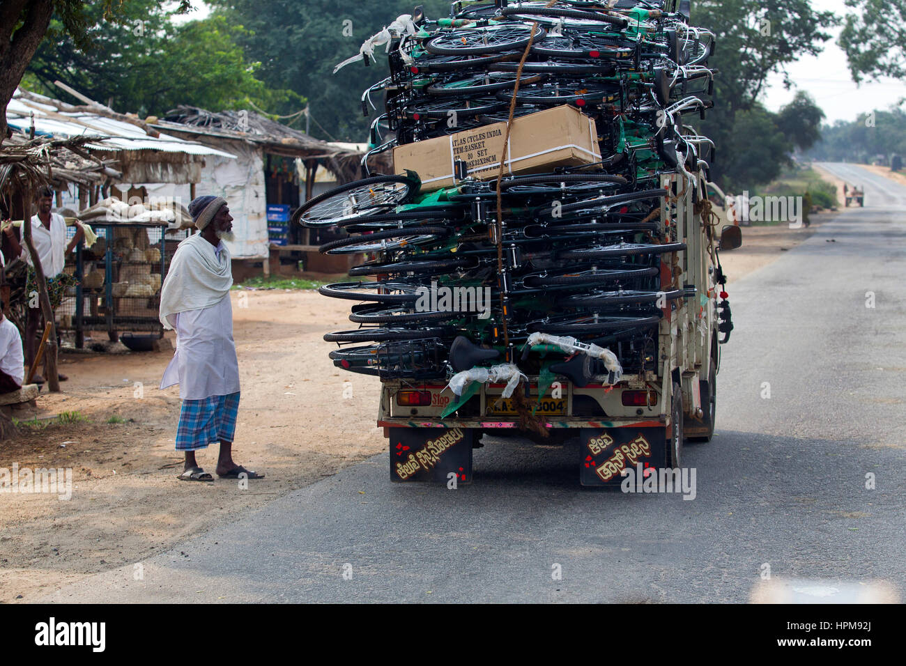 Indian man watching the passage of a small truck carrying dozens of bicycles in a Karnataka road, India Stock Photo