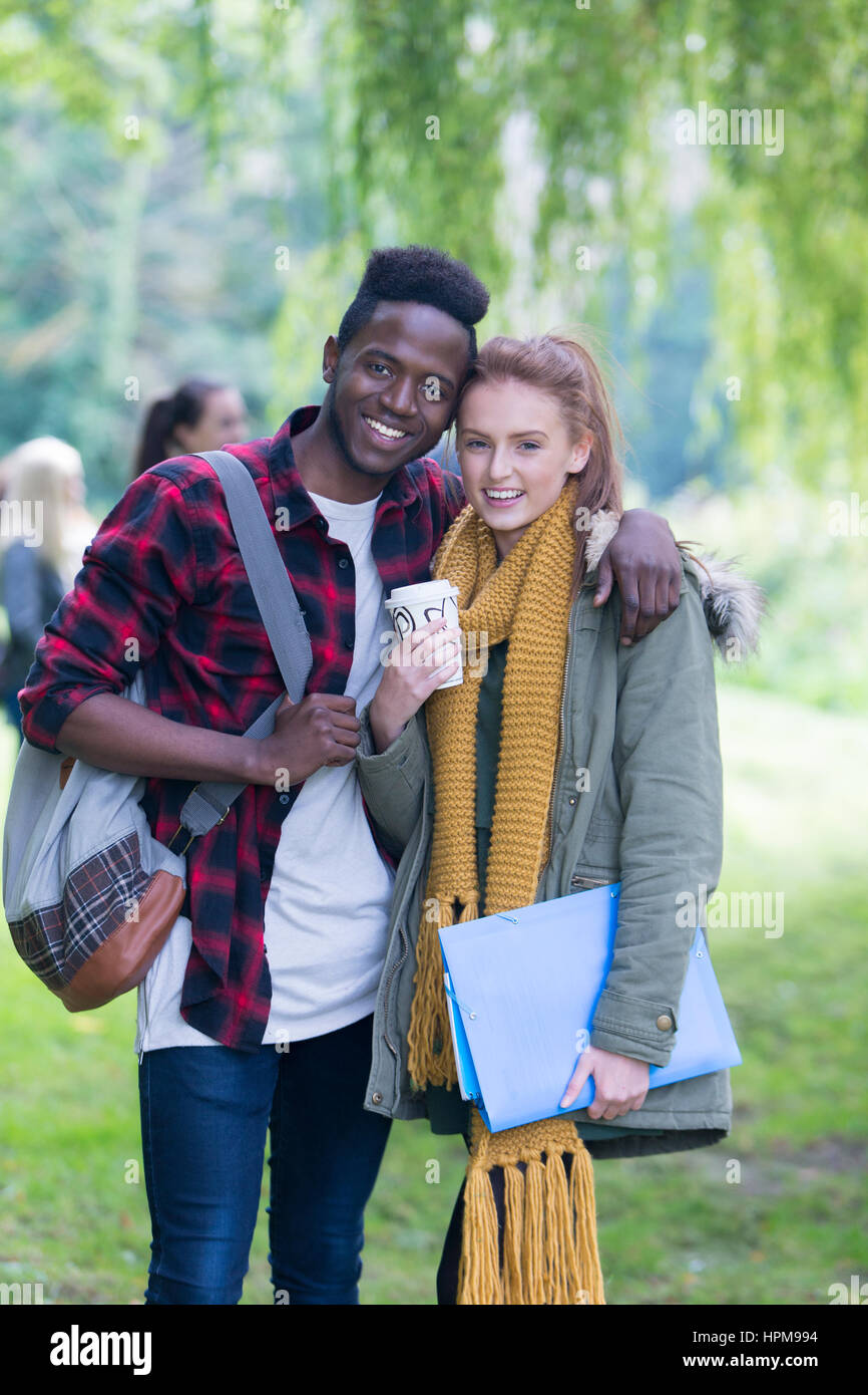 A happy young couple smiles as they relax outdoors with their friends. They are on a break from studying standing in an embrace and the girl is holdin Stock Photo