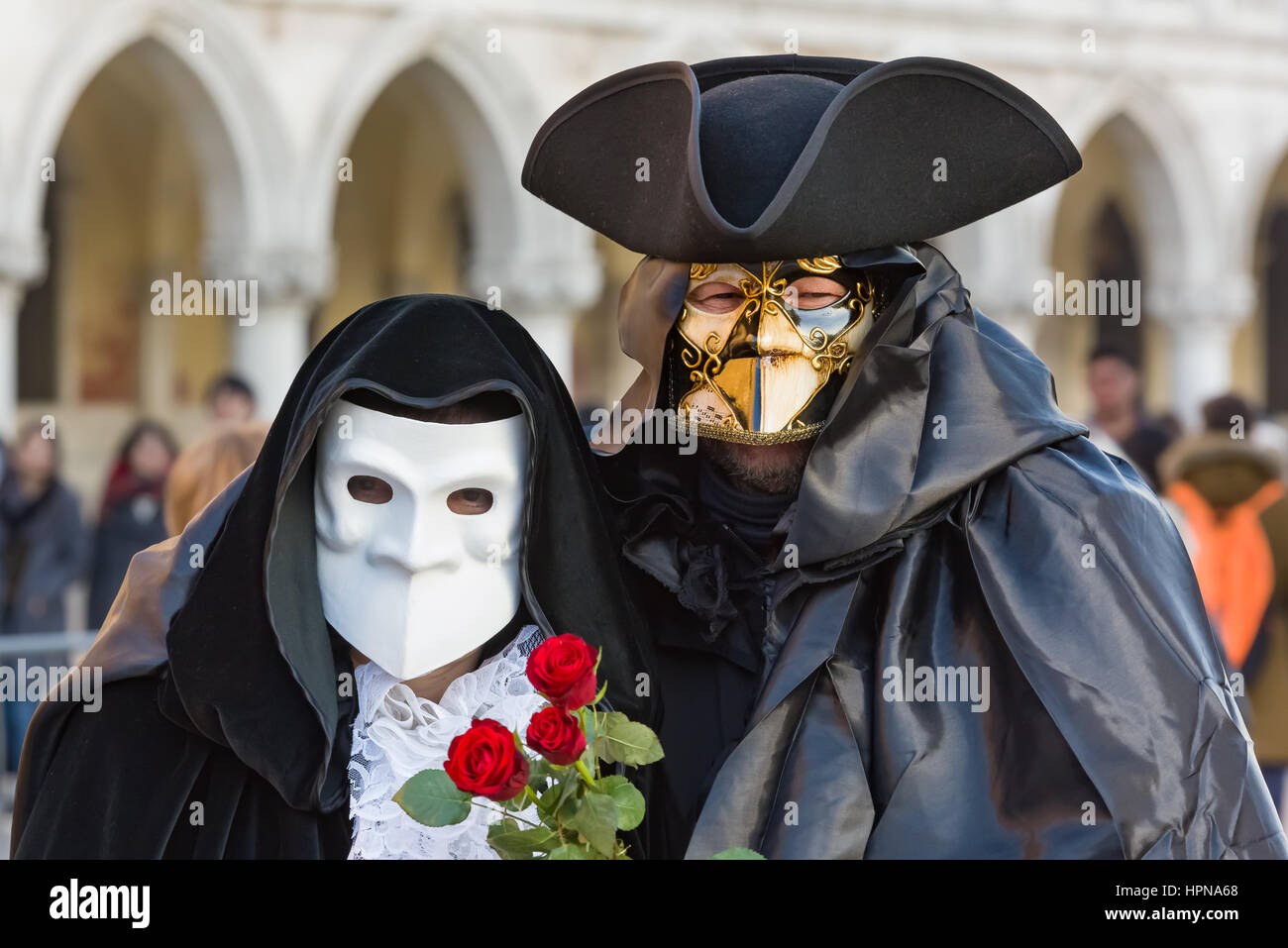 A couple are seen walking alongside the Doge's palace in St Mark's square Venice. they are wearing the traditional Bauta mask and cape, Stock Photo
