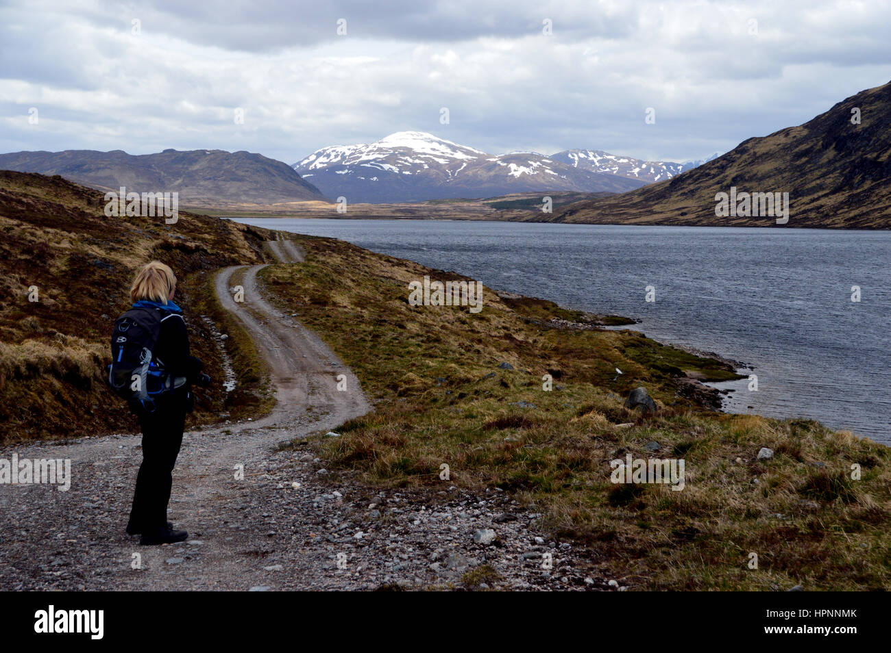 Lone Female Hiker Looking Down Lochan na h-Earba to the Snow Covered Scottish Mountain Munro Stob Coire na Ceannain in the Scottish Highlands, Stock Photo