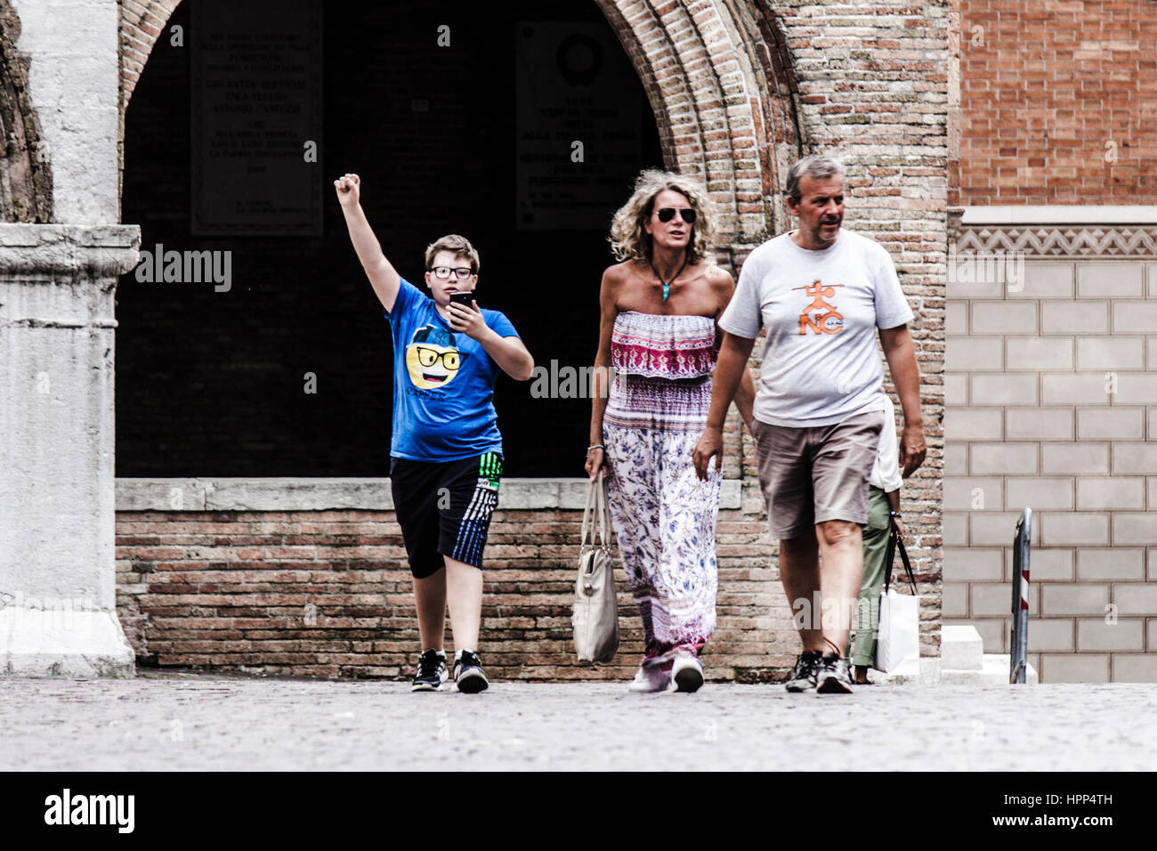 A family is walking on the street in Pordenone. Parents just walk; their son won some videogame using his phone Stock Photo